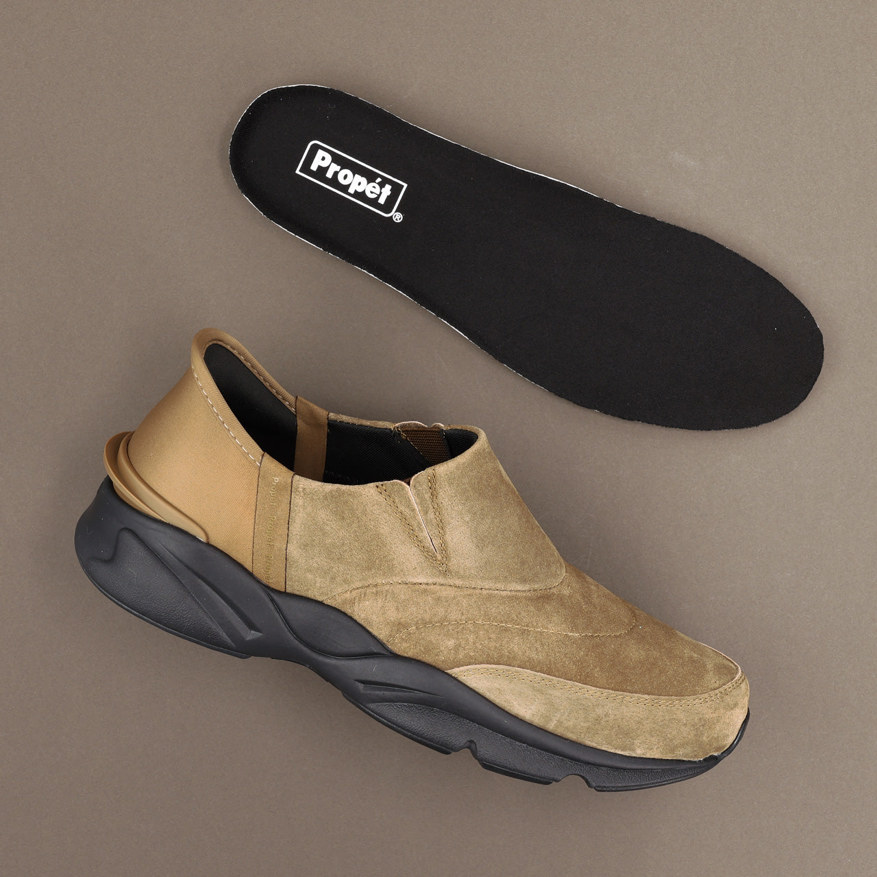 A tan and beige slip-on sneaker with a chunky black sole, shown alongside its black Propét branded insole on a neutral taupe background.