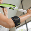 Lifestyle image of woman wearing the Actimove Sport Elbow Strap Hot/Cold Pack Universal while raising arm to swing tennis racket indoors.