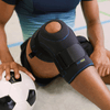 Lifestyle image of man wearing the Actimove Sport Knee Stabilizer Adjustable Horseshoe & Stays and sitting down on ground indoors while grabbing a soccer ball. Enjoy sports, as pain relief and healing are supported through balanced warmth and medical compression.