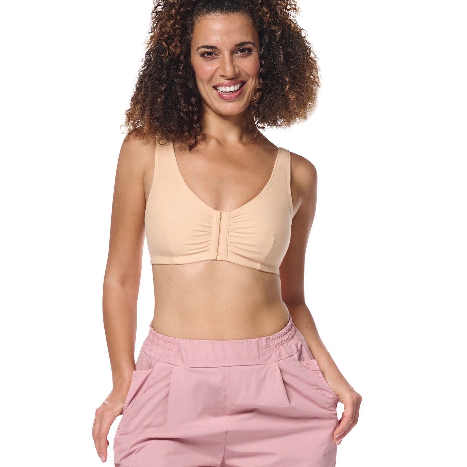 A smiling woman with curly brown hair wearing a beige ruched wireless bralette and dusty pink pants, photographed against a solid white background.