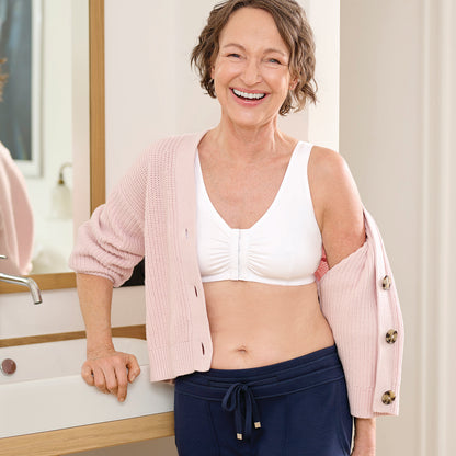 A smiling older woman stands indoors near a sink, wearing a light pink knit cardigan draped off her shoulders over a white bralette and navy drawstring pants.