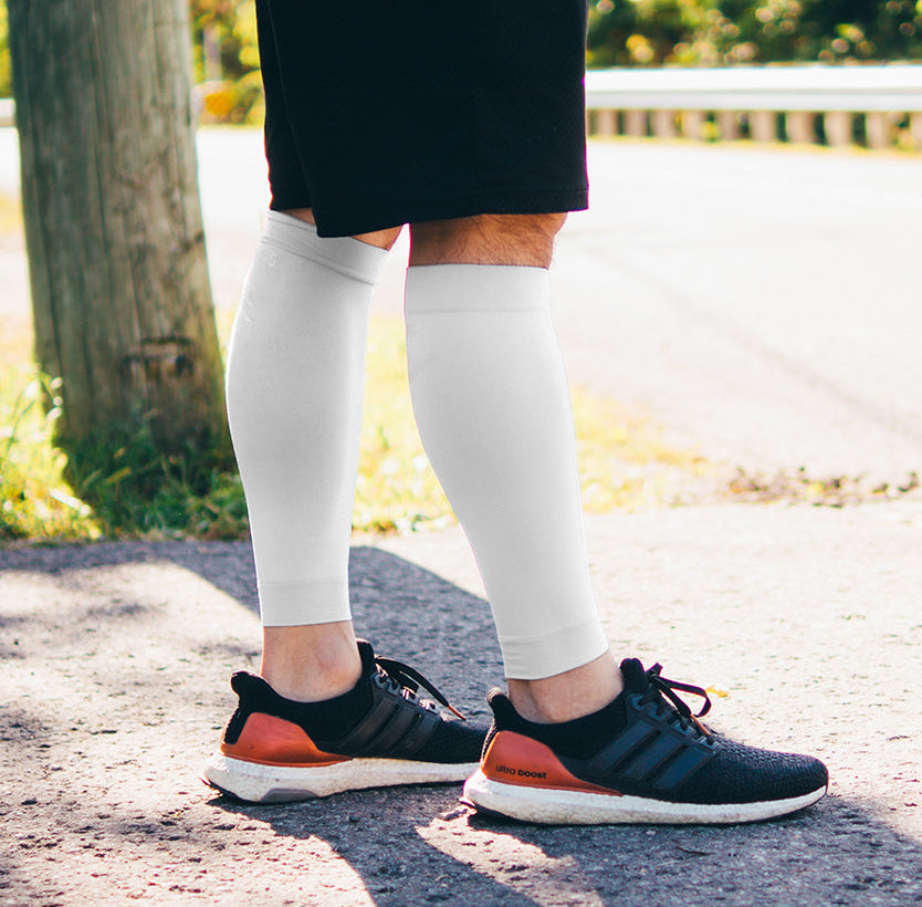 Person wearing white compression leggings and black and orange running shoes on a road with trees in the background, featuring the Sigvaris Group logo.