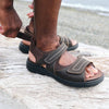 Close-up of man's feet wearing the sandals while adjusting the flexible back hook and loop strap for comfort and personalized fit