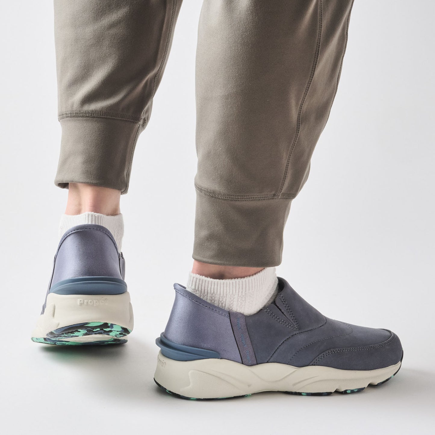 A person wearing gray jogger pants and white socks, shown from the calves down, wearing a pair of slip-on sneakers with a slate blue upper and chunky off-white midsoles.