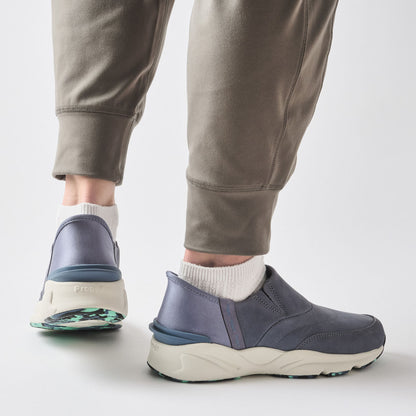 A person wearing gray jogger pants and white socks, shown from the calves down, wearing a pair of slip-on sneakers with a slate blue upper and chunky off-white midsoles.