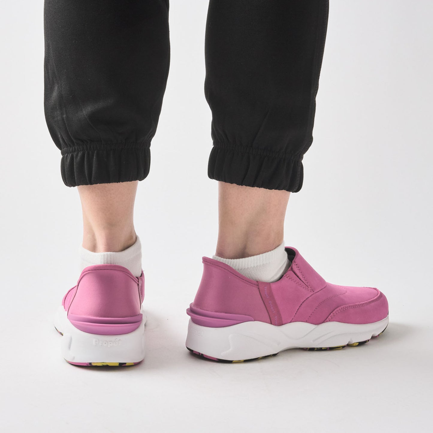 A person wearing pink slip-on sneakers with white soles, paired with black jogger pants and white socks, shown from the back against a light gray studio background.