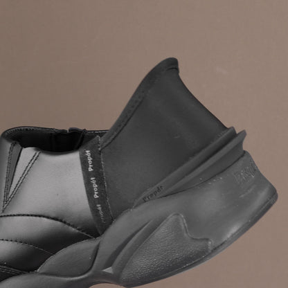 A close-up of a black slip-on shoe with a chunky sculpted sole, featuring a side strap with 'Project Project' text, set against a solid taupe background.