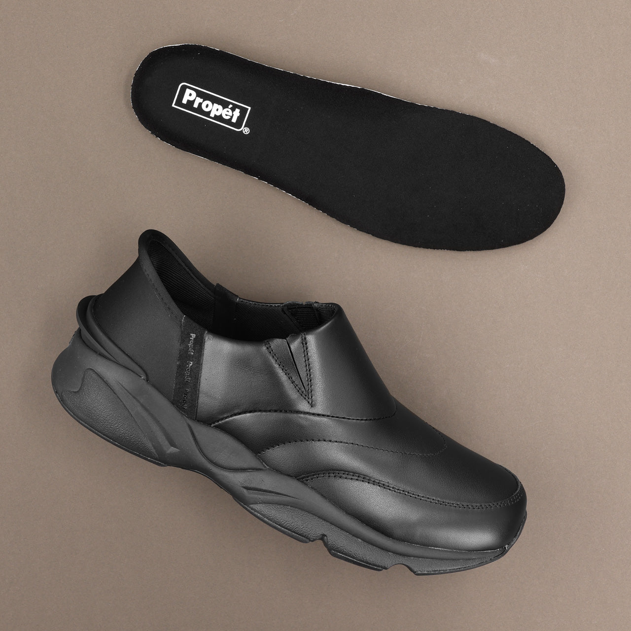 A black slip-on shoe shown from the side with its matching black insole placed above it on a plain taupe background. The insole has a visible white 'Propél' logo.