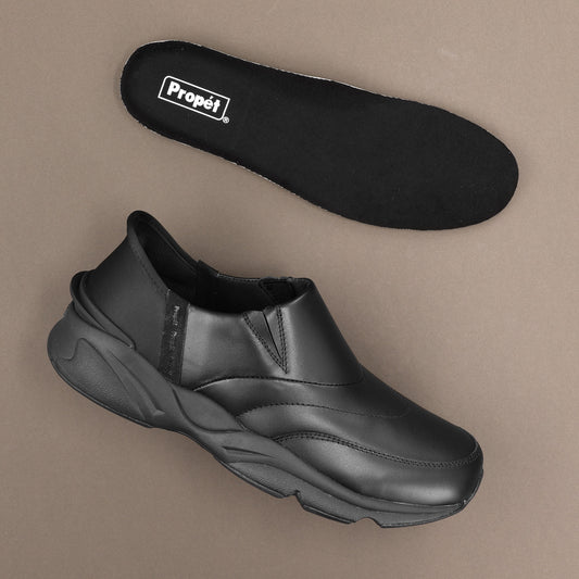 Propet Men's Stability Freestyle Step In Sneaker - A black slip-on shoe shown from the side with its matching black insole placed above it on a plain taupe background. The insole has a visible white 'Propél' logo.