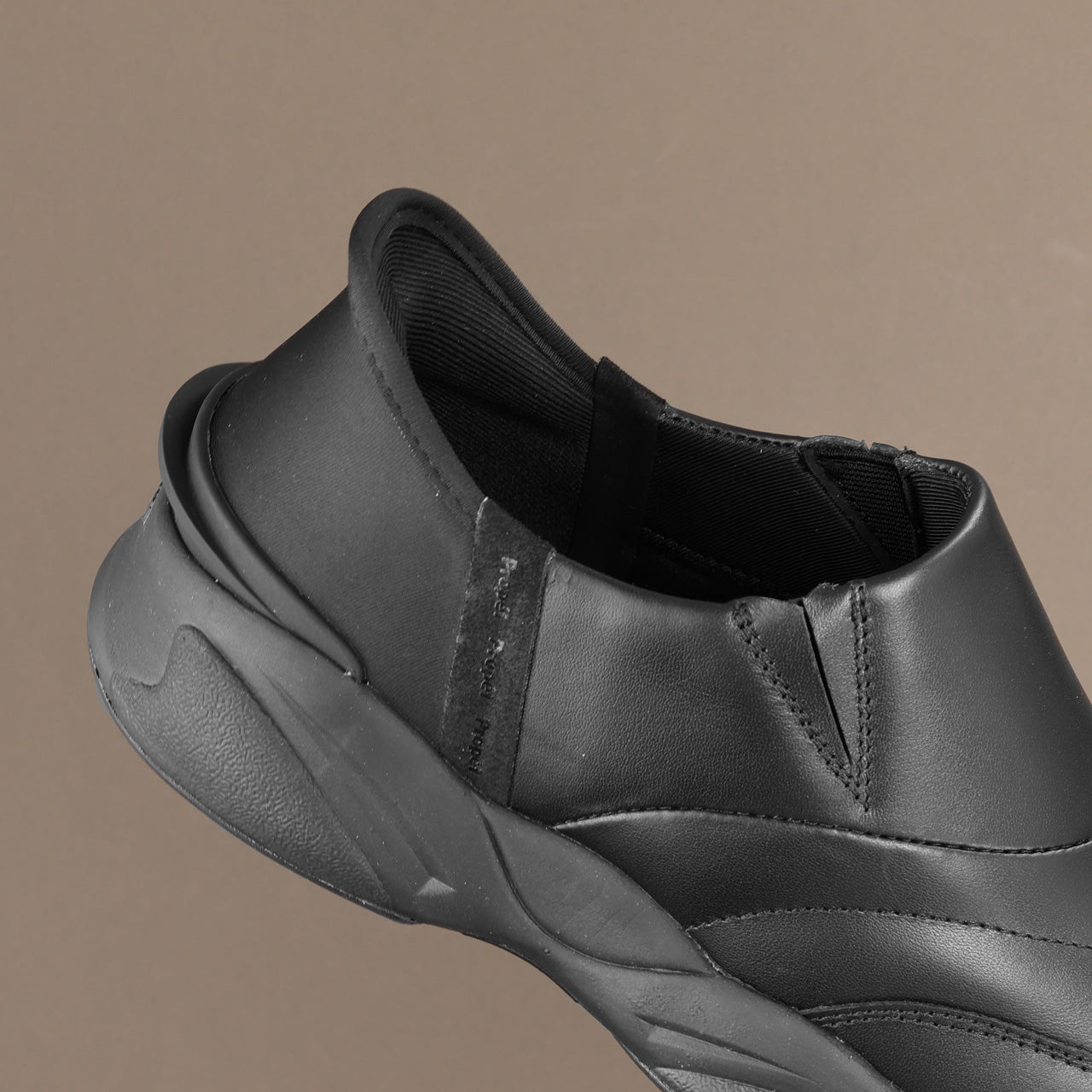 A close-up of a black slip-on sneaker showing the side profile, heel, and textured sole against a solid taupe background.