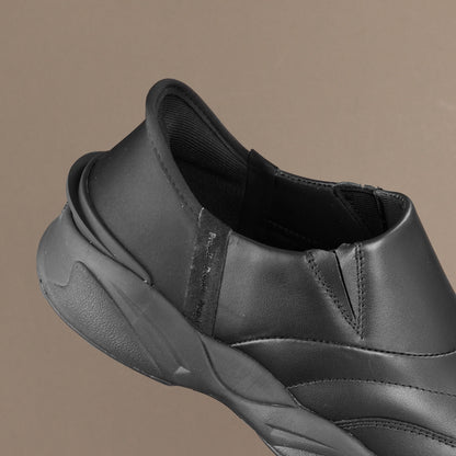 A close-up of a black slip-on sneaker showing the side profile, heel, and textured sole against a solid taupe background.