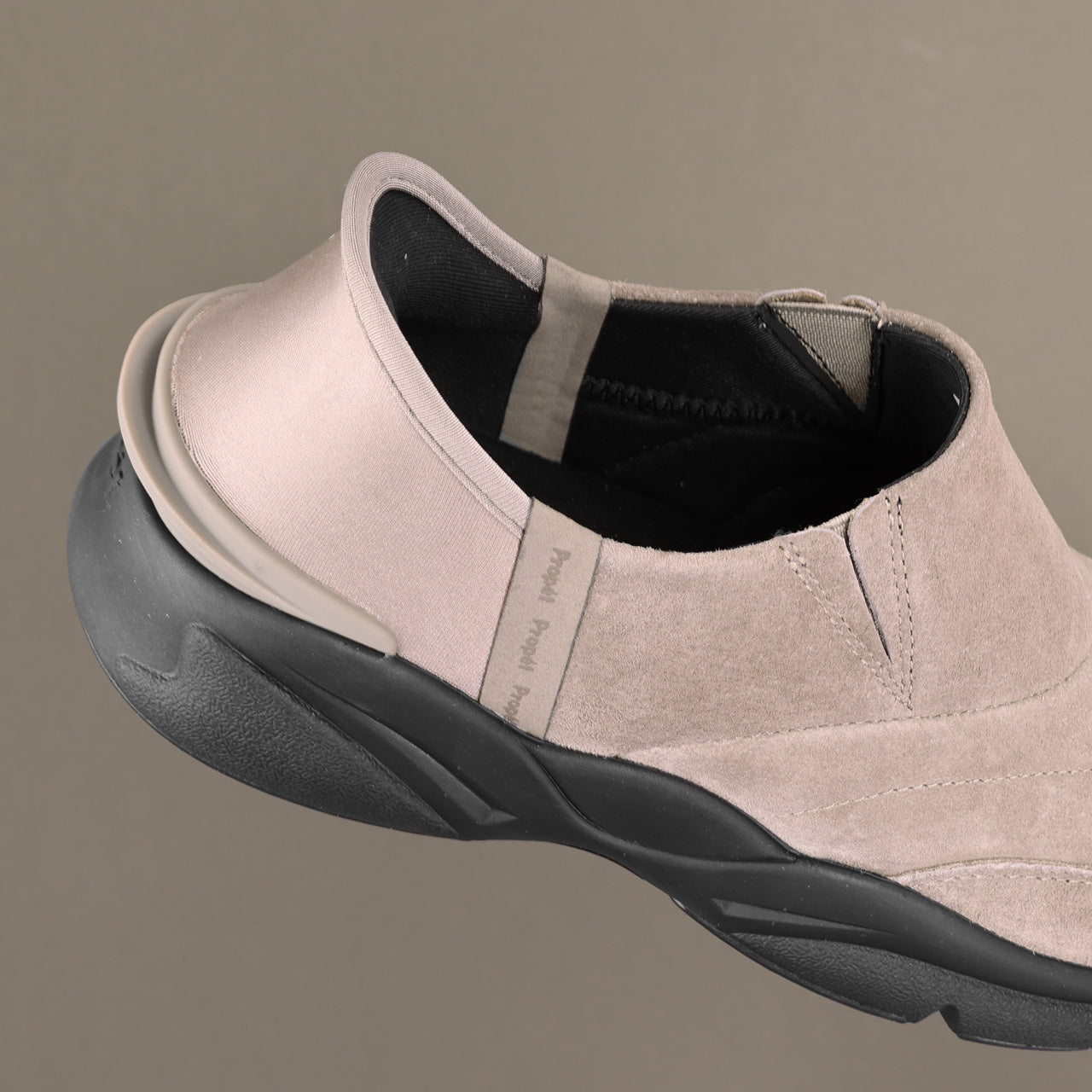 A close-up side view of a slip-on shoe with a light beige suede upper and a thick black sole, shown against a solid taupe background. The brand name 'Paul Reed' is visible on a small fabric tag.