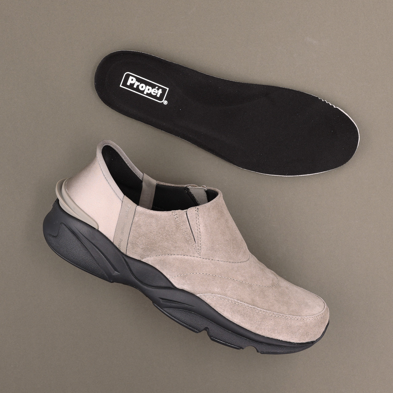 A light beige slip-on sneaker with a chunky black sole, shown alongside its black Propét insole on a neutral taupe background.