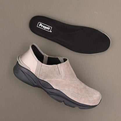 A light beige slip-on sneaker with a chunky black sole, shown alongside its black Propét insole on a neutral taupe background.