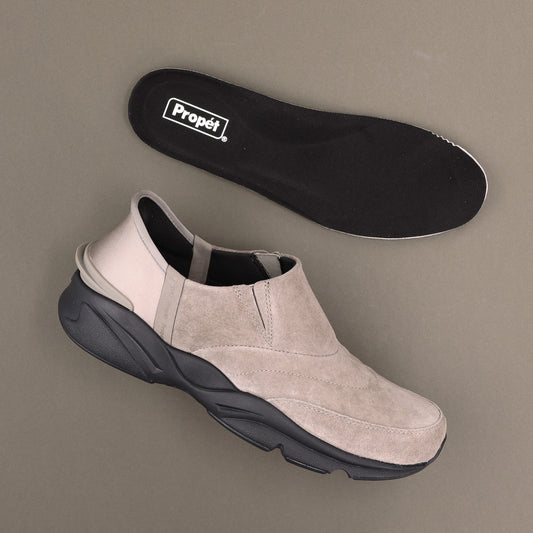 Propet Men's Stability Freestyle Step In Sneaker - A light beige slip-on sneaker with a chunky black sole, shown alongside its black Propét insole on a neutral taupe background.
