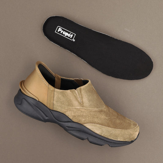 Propet Men's Stability Freestyle Step In Sneaker - A tan and beige slip-on sneaker with a chunky black sole, shown alongside its black Propét branded insole on a neutral taupe background.