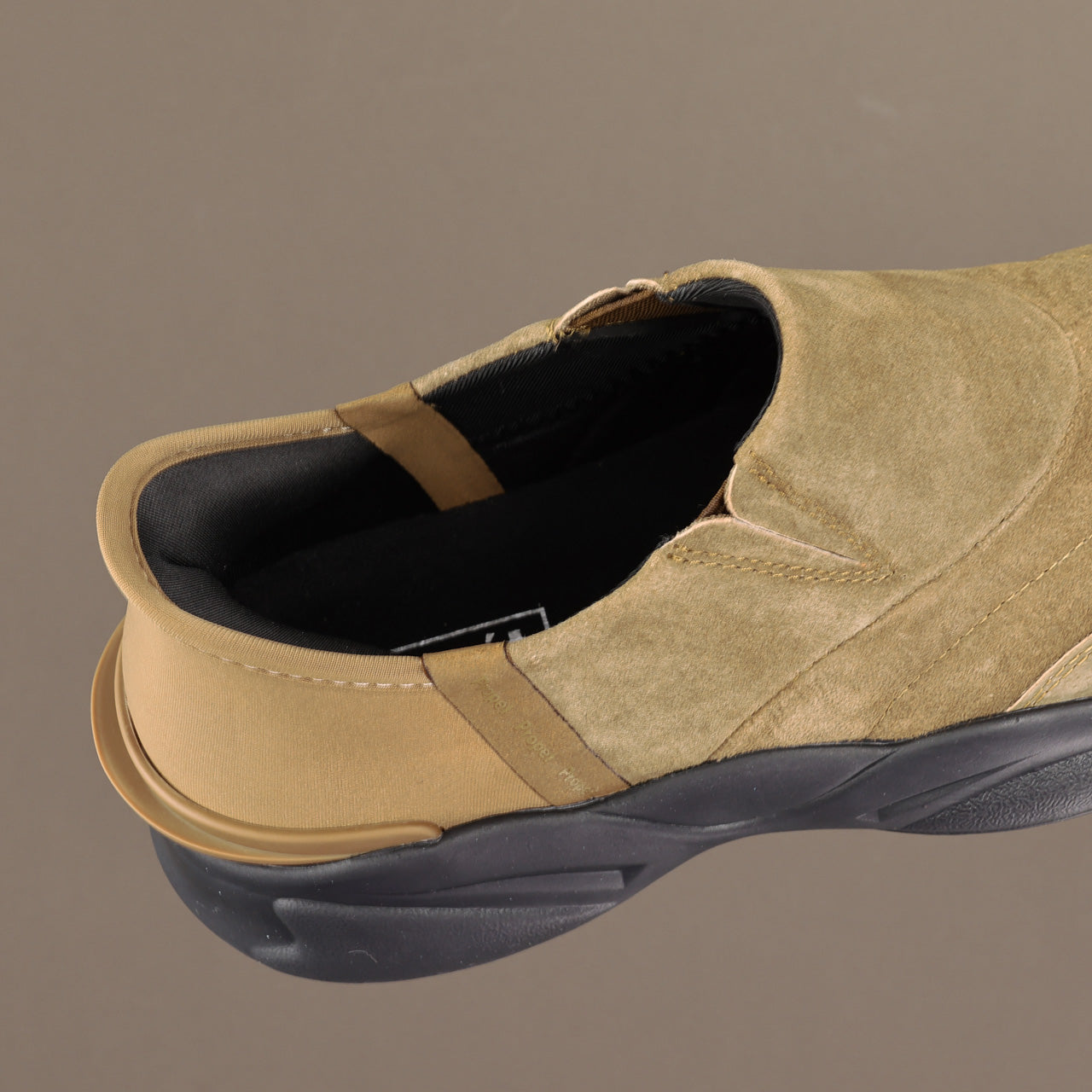 A close-up side view of a slip-on shoe in tan suede with a black sole and black interior lining, set against a solid taupe background.